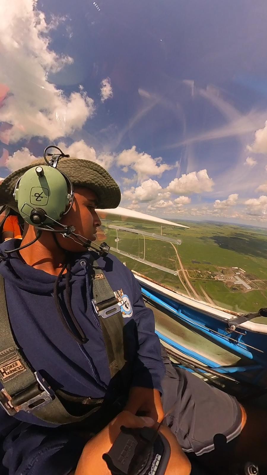 View from the front seat in flight: a student in a headset looks out over green prairie fields and the airfield below