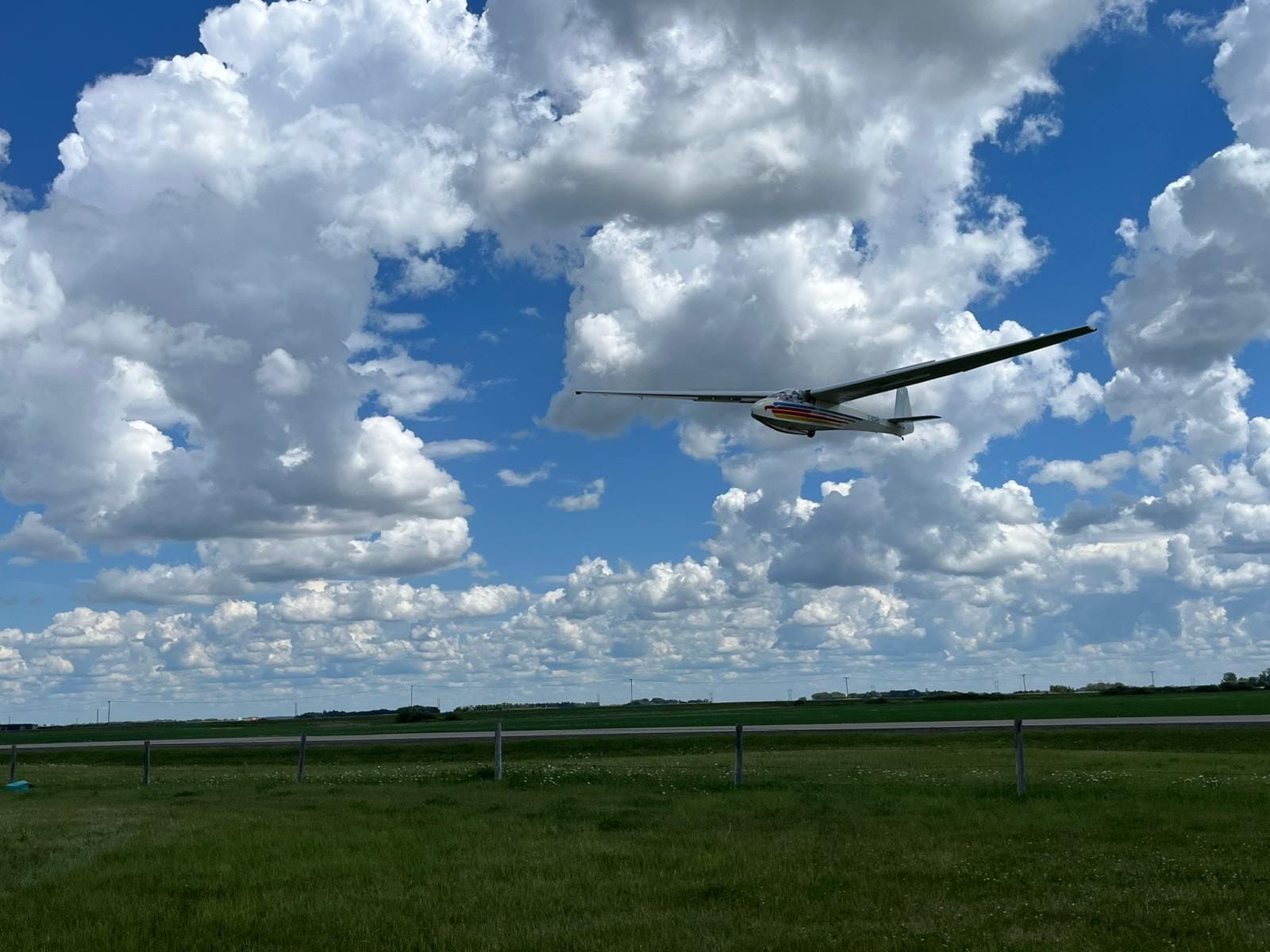 A club glider flies low over the field against towering white cumulus clouds