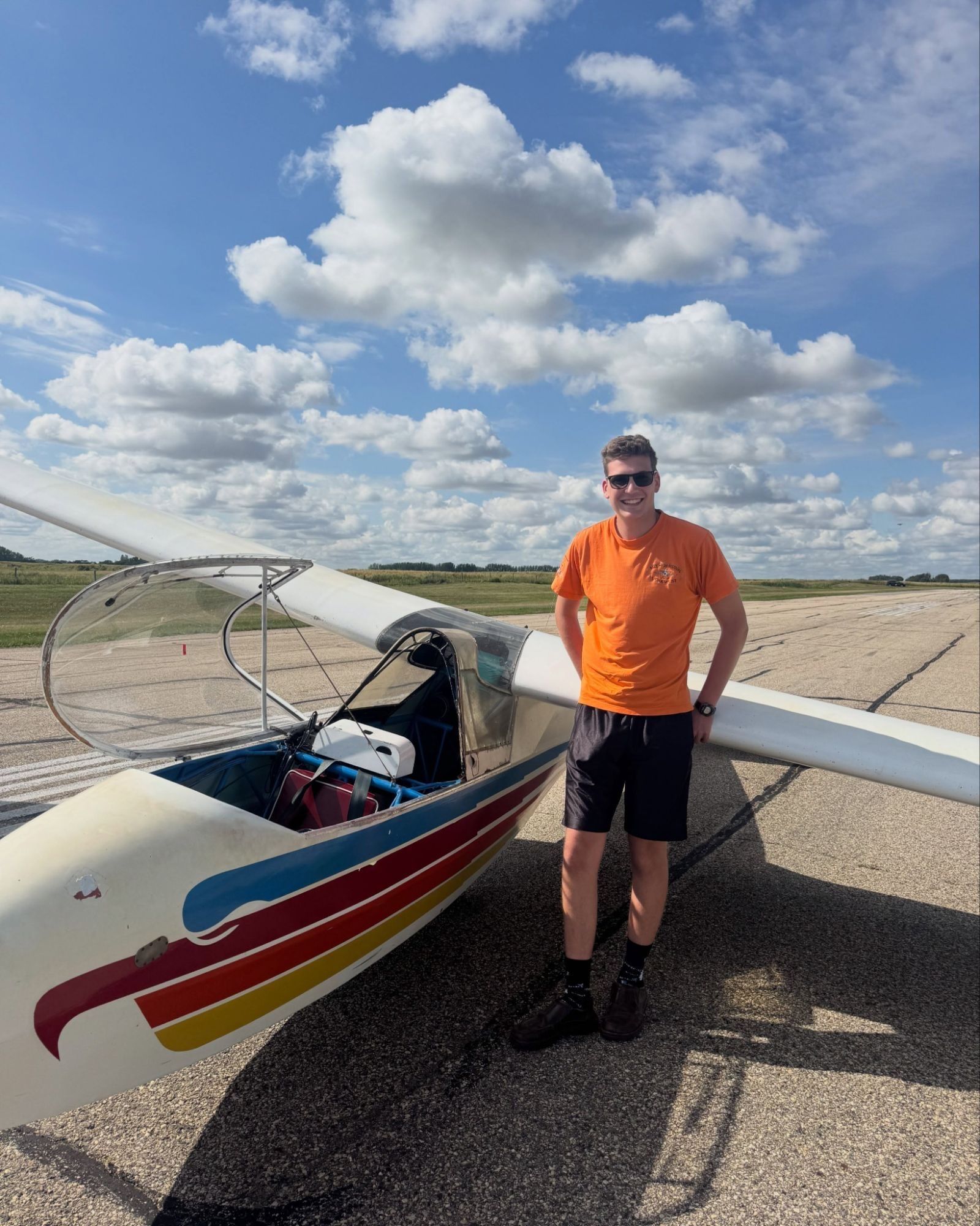 A smiling student stands beside the club's two-seat glider on the runway under a sky full of cumulus clouds